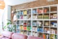 Bookshelf with color-coordinated books, pink sofa, potted plants, and a round mirror on a brick wall.