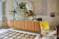Mid-century modern living room with a wooden sideboard, round mirror, plants, wicker chair, and geometric rug.