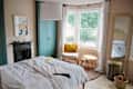 Bedroom with teal wardrobe, fireplace, and bay window. Features a bed, chair with yellow cushion, round tables, and a large mirror.