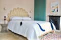 Bedroom with a rattan headboard, white quilt, teal wardrobe, abstract art, and a colorful round rug.