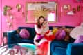 portrait of woman holding fluffy cat on sofa. Bright pink wall and lots of artwork, mirror, gold accents, behind