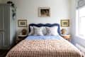 Blue upholstered scalloped headboard in bedroom with neatly made bed with brown and white gingham quilt. Small portraits hung around bed with   straw pendant hung above bed.