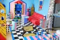 Living room with bright blue walls with darker blue trim, wood floor with black and white checkered tile "runner", red sofa, lots of small tables and pedestals in geometric shapes in many different colors, pink and blue striped dining table peeking in the front of the frame with pink and blue wavy candlesticks, other colorful decor.