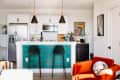 Modern kitchen with teal bar, two green stools, orange armchair, pendant lights, and stainless steel appliances.