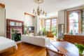 Bright living room with white sofa, wooden floors, fireplace, chandelier, and large windows with plants and decor.