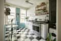 kitchen with white cabinets, black and white checkered floor, white tile backsplash, blue walls with white trim. Shelves over stove area with spices, pans. Plant leaves in white pitcher on counter. To the left is the dining area with small round marble bistro table with clear lucite chairs. Black and white checkered floor, blue walls and radiator with white trim. Pots and pans hanging above dining table. 2 windows letting in light