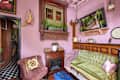 living room with pink walls, pink and white decorative ceiling, green velvet antique sofa with shiny pink velvet pillow and throw, tufted rose velvet armchair next to small wood fireplace, wood floors, lots of shelves, doorways, windows with detailed wood trim, plants, art objects