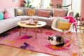 pink living room with pastel lilac sofa with colorful pastel pillows. Pink shag area rug with hot pink, purple and yellow design, round light wood coffee table, white modern rocking chair with gray and yellow throw pillows, bright pink throw, large windows, framed art, potted trees on floor and hanging plants in window. Cat on rug