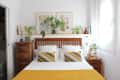 bedroom with white walls, bed with mustard, gray and white bedding with wood headboard, wood side tables, shelf over bed with plants, art