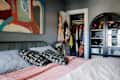 bedroom with gray walls, large abstract painting over bed, black metal headboard, bed with black and white patterned throw pillows, mushroom shaped metal lamp on bedside table, closet open in background with clothes hanging, black and glass arched cabinet with accessories
