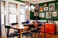 Dining room with green walls with white trim, wood floors, gallery wall, wood dining table with black and gray patterned chairs, "faux" tiger rug, stack of books on floor, orange cabinet with bar tray setup