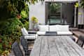 Plant filled outdoor space with wooden dining table and separate lounge area with wrought iron seating topped with white cushions.