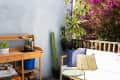 sunny outdoor patio area with wood bench with off white cushions, small green table, outdoor rug/mat, blooming bougainvillea plants and other potted plants