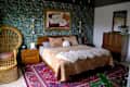 Bedroom with green floral wallpaper and wicker chair in corner beside bed. Bed is made with tan velvet blanket and animal print linens. Vintage wooden dresser across from bed.