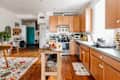 Spacious kitchen with wooden cabinets, island with fruit, colorful rug, and teal door in the background.