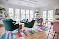 Open, sunny dining room with two seating areas. One round dining table surrounded by printed dining chairs and another round conversation area. Colorful rug lines the floor.