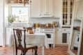 A white dining room table for two near a kitchen with all white cabinets and appliances on a tile floor.