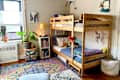 Cozy children's bedroom featuring a wooden bunk bed, colorful rug, bookshelves, and plants. Decor includes toys and artwork.
