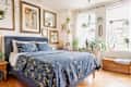 Cozy bedroom featuring a blue floral bedspread, framed art on the walls, and various indoor plants by the windows.