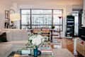 Cozy living room with a light sofa, glass coffee table, and dining area featuring rattan chairs and a view of the city.