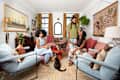 Three people in a cozy living room with plants, cats, and colorful decor, sitting on pink sofas and laughing.