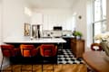 Modern kitchen with white cabinets, black and white checkered floor, orange velvet bar stools, and wooden dining table.