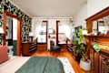 Cozy bedroom featuring a green blanket on a bed, wooden furniture, large windows with sheer curtains, and decorative plants.