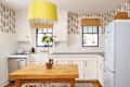 Bright kitchen with white cabinets, blue windows, a yellow pendant light, and a wooden table with a potted plant.
