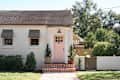 Charming house exterior with a pink front door, gray awnings, potted plants, and a brick step leading to a garden gate.