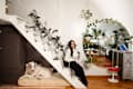 A woman sitting on white stairs with plants, a large dog on a bed below, and a round mirror reflecting a cozy living space.