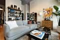 Marble coffee table and gray sofa in front of shelving unit across from large floor mirror, wall sconce, art, bar cabinet, and potted tree.