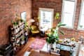 Cozy living space with exposed brick walls, yellow armchair, bookshelves, plants, and a colorful rug.