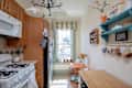 Cozy kitchen with wooden cabinets, white stove, orange table, floral cushions, and hanging mugs on a blue shelf.