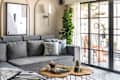 Cozy living room with a gray sectional sofa, decorative pillows, wooden coffee table, and large plants near glass doors.