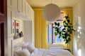 Cozy bedroom with a white bed, yellow curtains, a large plant, and decorative items on shelves.