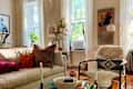 Cozy living room with a light sofa, colorful pillows, a vintage bullfighting poster, and plants by the window.