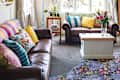 Bright living room with colorful pillows on a brown leather sofa, floral rug, and a wooden coffee table with flowers.