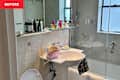 Cluttered bathroom sink area with various skincare products, a pink cosmetic bag, and a shower stall in the background.