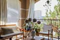 Cozy balcony with wooden furniture, potted plants, and large purple flowers in a vase, surrounded by sheer curtains.