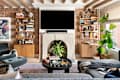 Cozy living room with exposed brick walls, a large TV above a stone fireplace, bookshelves, and a green plant.