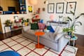 Cozy living room with a gray sofa adorned with colorful pillows, an orange side table, and plants. Wall art and a patterned rug enhance the decor.