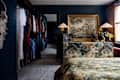 A cozy bedroom featuring a floral bedspread, a decorative dresser, a painting, and a closet with colorful clothing.