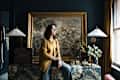 A woman in a gold top sits on a colorful dresser, with a vintage painting behind her, a lamp, and plants nearby.