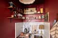 Cozy kitchen corner with red walls, wooden shelves displaying glassware and pottery, and a small sink with decor.