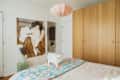 A white cat standing on a bed with a colorful quilt, wooden wardrobe, and a large art piece on the wall.