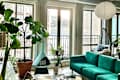 Bright living room with large windows, green velvet sofa, hanging chair, and various indoor plants.