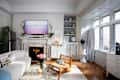 Bright living room featuring a white sofa, wooden chair, floral decor, and a marble fireplace with a TV above.