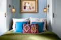 Cozy bedroom with a green blanket, blue and patterned pillows, and woven wall sconces. Framed art above the bed.