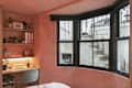 Cozy bedroom corner with pink walls, a desk with a plant, books, and a chair, featuring large windows overlooking stairs.