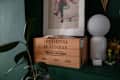 Wooden crate labeled "LES JARDINS DE LEOGNAN" beside a white lamp, framed artwork, and a potted plant on a green wall.